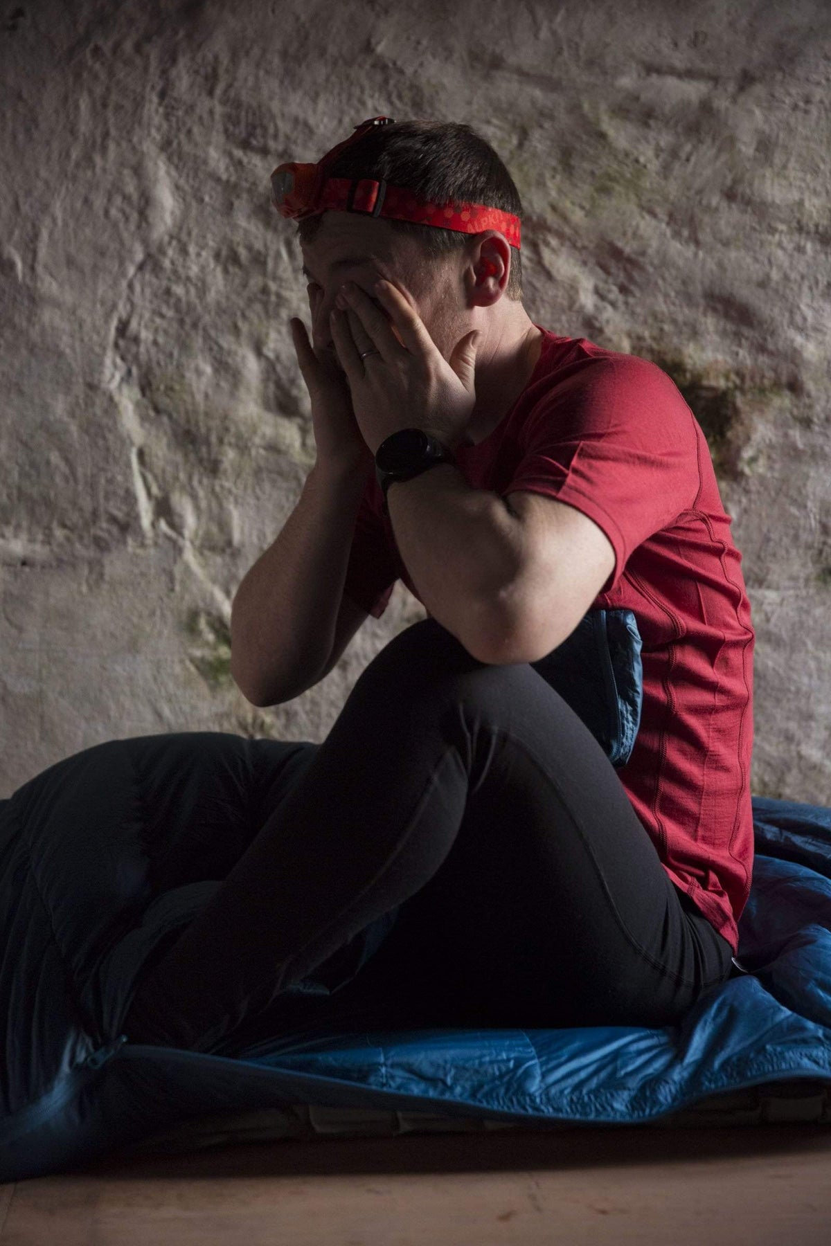 A man waking up in his sleeping bag, rubbing his eyes in a merino wool top and a head torch on his head - action